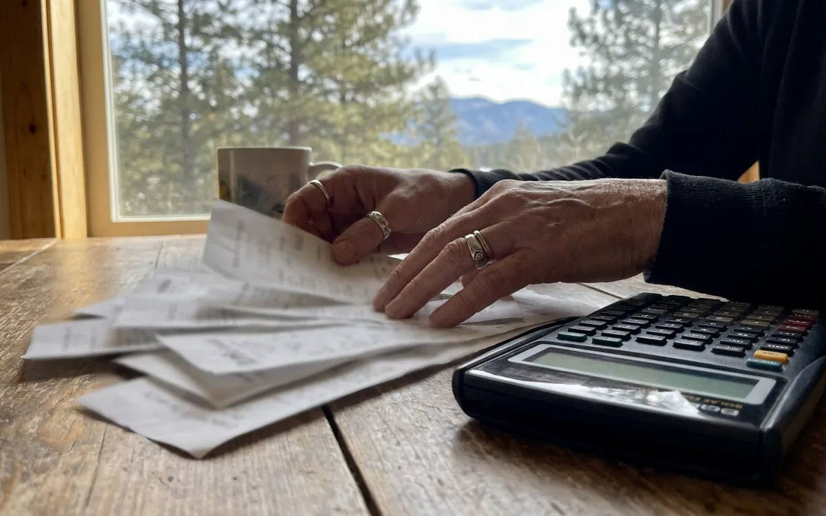 Close-up of hands sorting paper invoices next to a calculator on a wooden desk near a window