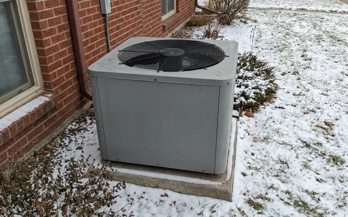 Outdoor heat pump unit on a raised pad beside a brick house with light snow on the ground